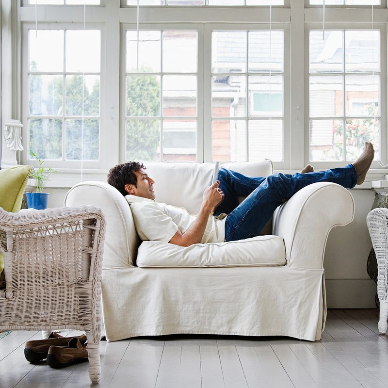 Man laying down on couch reading book next to window.