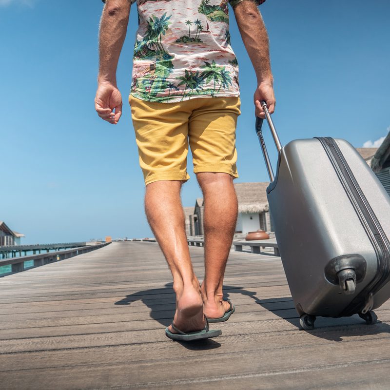 A man wearing a Hawaiian shirt and yellow shorts is seen pulling his luggage to his hotel room in a tropical destination.