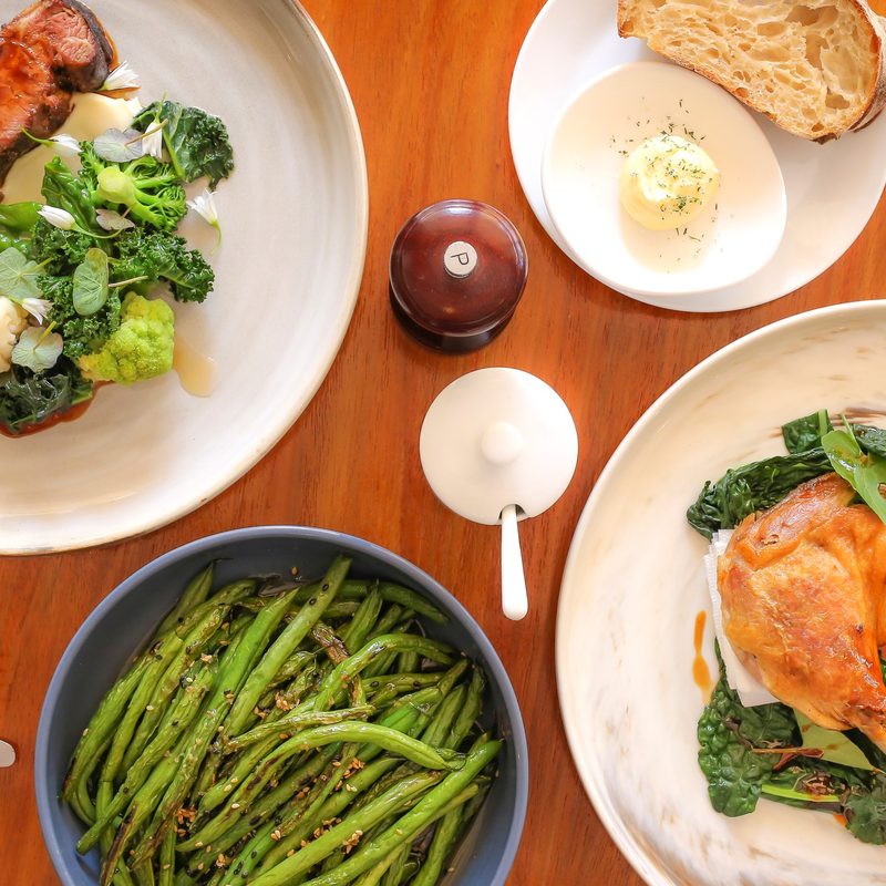 Chicken and steak meals on the table with salt and pepper as well as veggies, green beans, sourdough bread, and butter on the side.