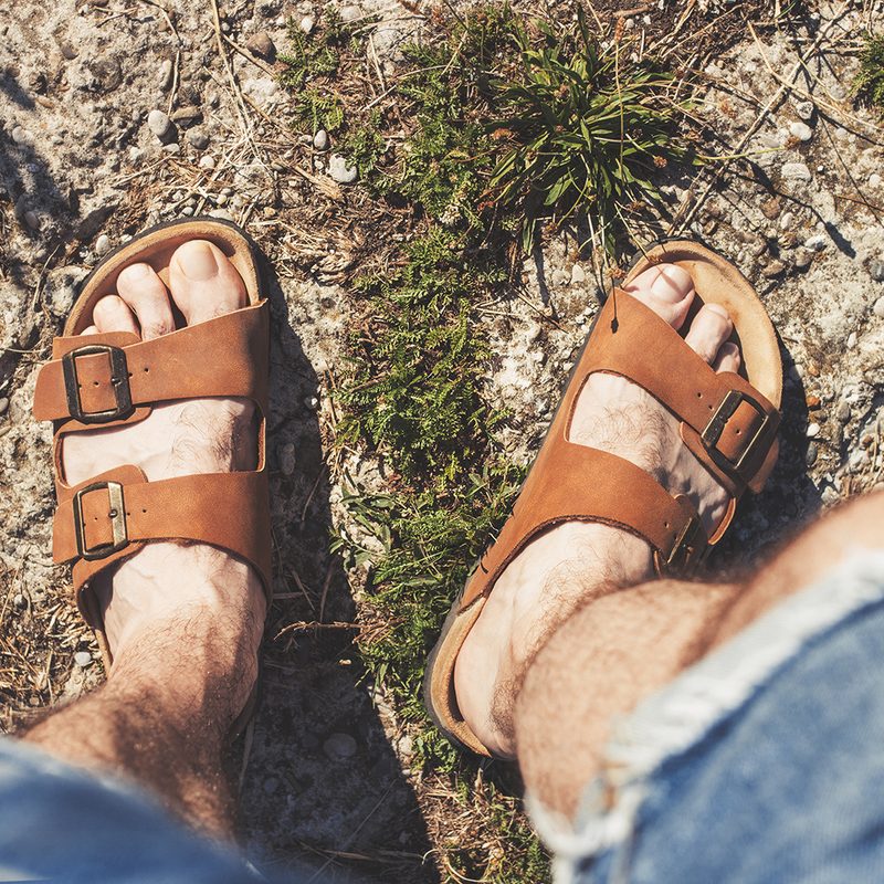 Downward view of a man's feet wearing brown sandals standing on grass.