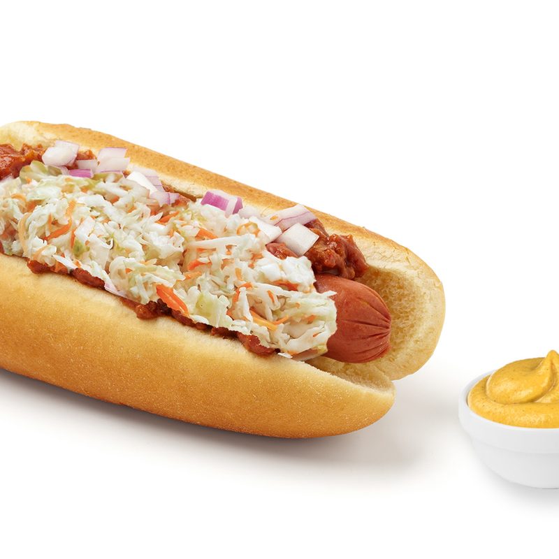 A hot dog with coleslaw, onions, and chili next to mustard dressing on a white background