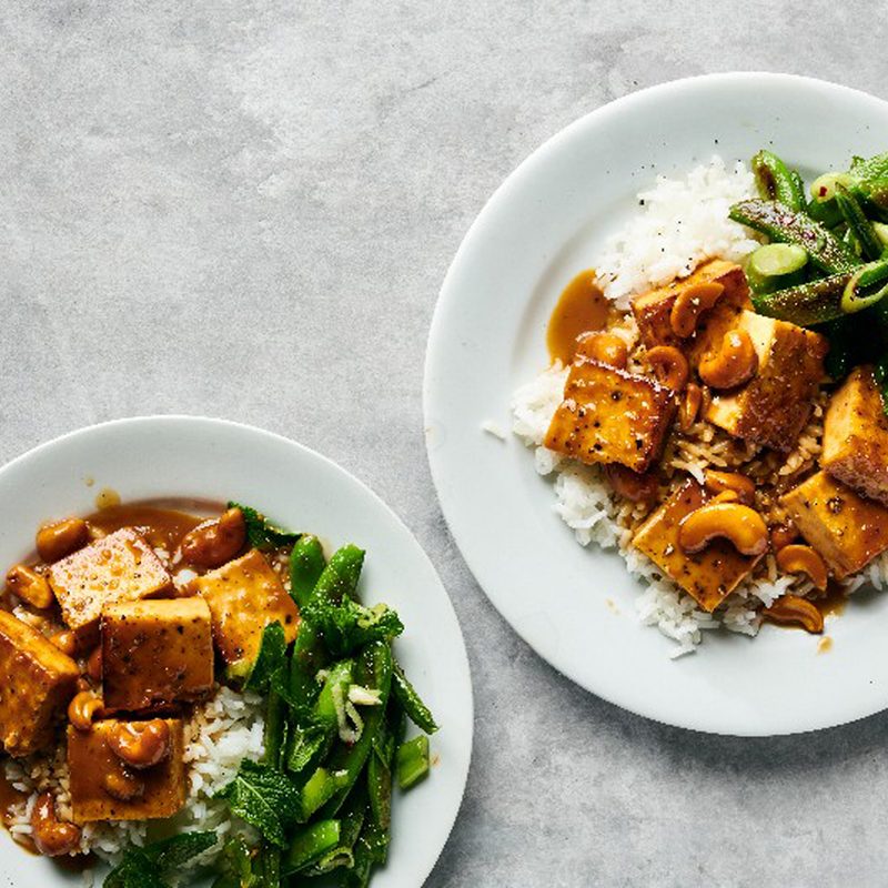 Two plates of crispy tofu with cashews and blistered snap peas on a table.