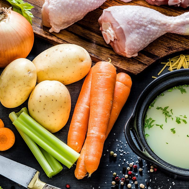 Raw chicken thighs on a chopping board, carrots, potatoes, noodles, olive oil, onions, celery, parsley, and a bowl of broth.
