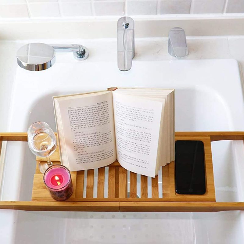 A Dozyant Bamboo Bathtub Tray Caddy with wine glass, candle, an opened book, and phone on top of a sink in a bathroom.