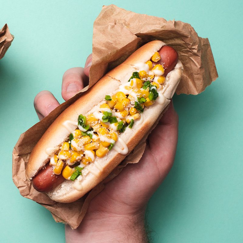 Hand holding Mexican street style corn cilantro and cheese on hotdog on a teal background