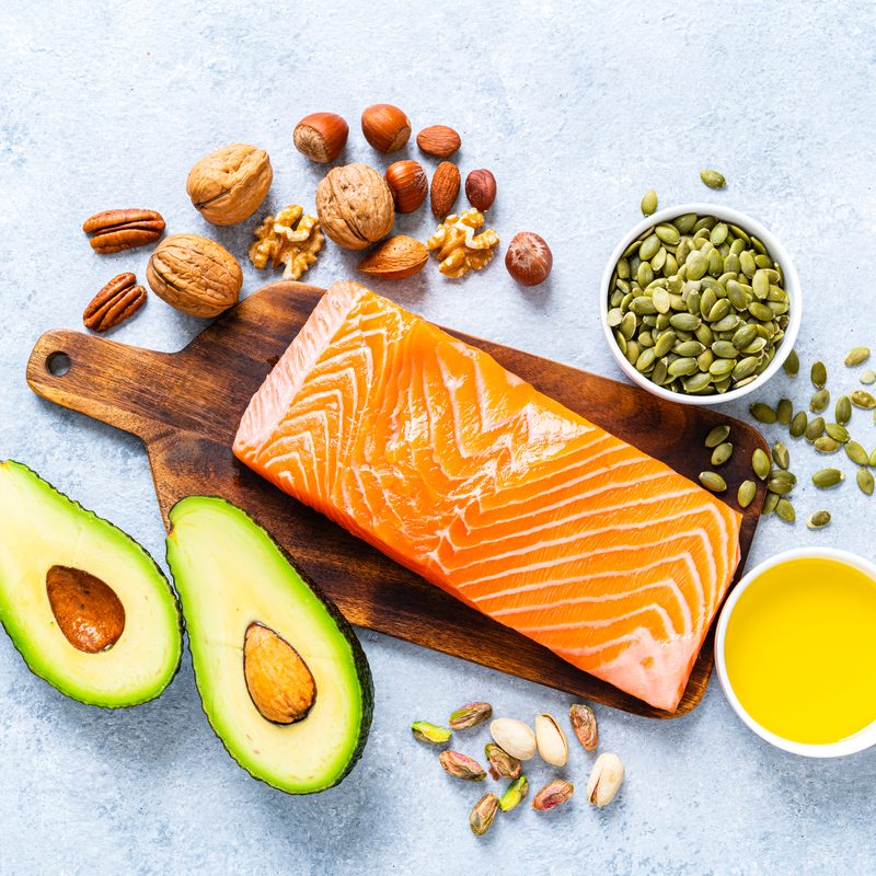 An overhead shot of salmon on cutting board, avocado, extra virgin olive oil, nuts, and bowl of seeds on a table