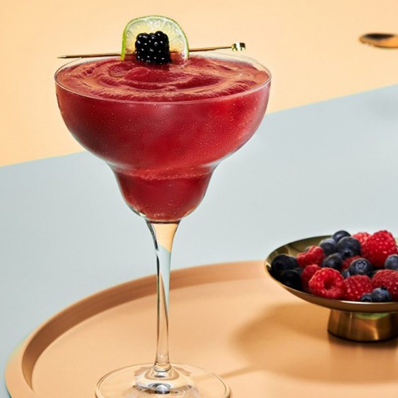 A glass of Frozen Berry Margarita beside a bowl of berries on a circular tray.