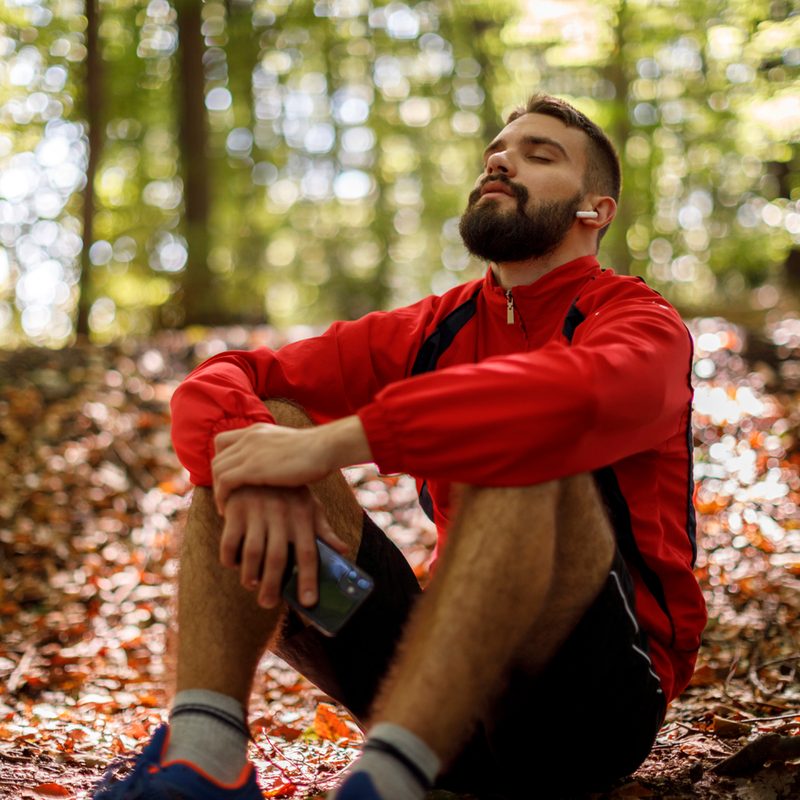 A man sitting and listening to music through his Bluetooth earbuds in the forest.