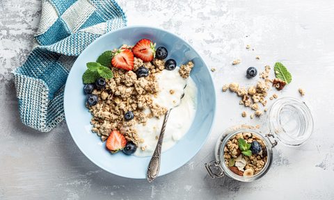 A blue towel, a bowl of yogurt with granola, blueberries, strawberries, and a mint leaf, and a jar of granola on a table.