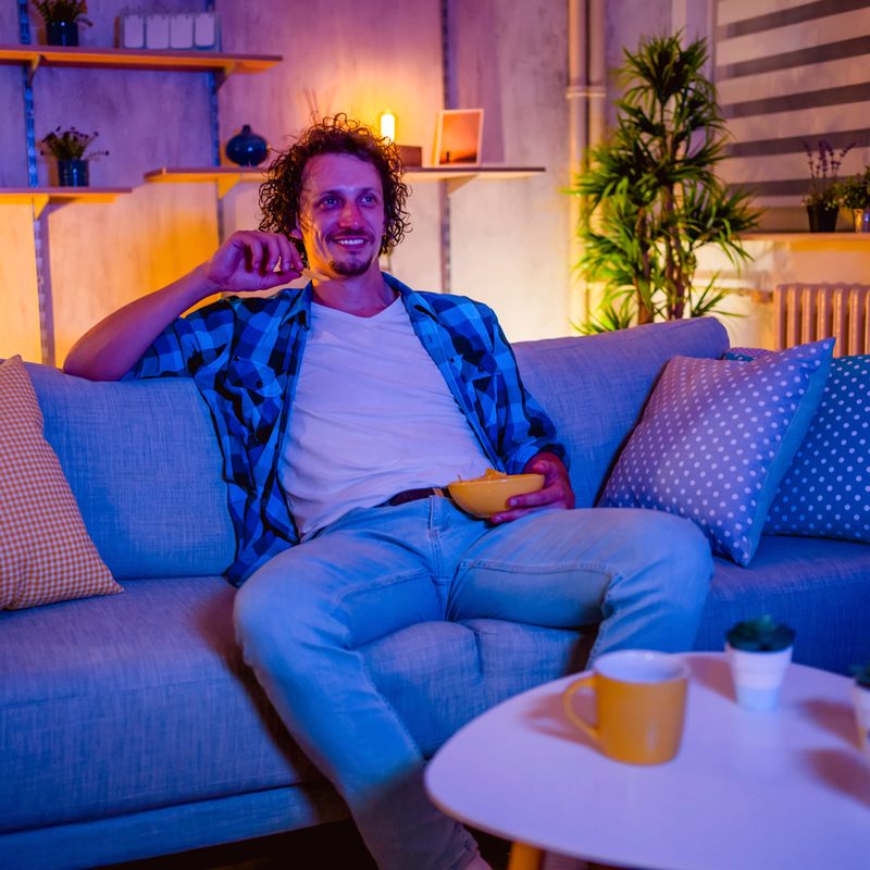 A smiling man watching TV and eating snacks in the living room.