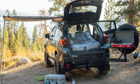 A parked vehicle with a blanket and Luno air mattress, camping stove, camping lantern, and cooler in a campsite.