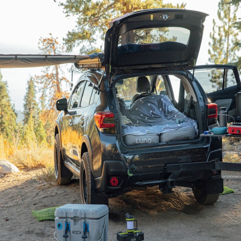 A vehicle with a blanket and Luno air mattress, camping stove, camping lantern, and cooler in a campsite.