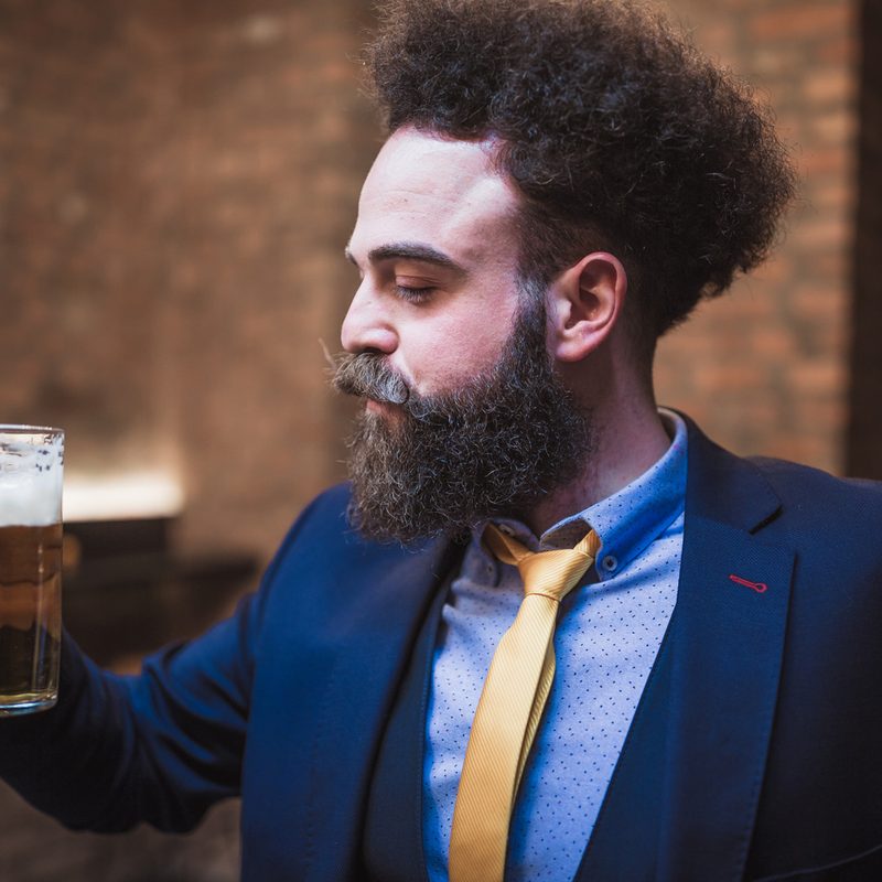 A bearded man in a suit holding a glass of beer.