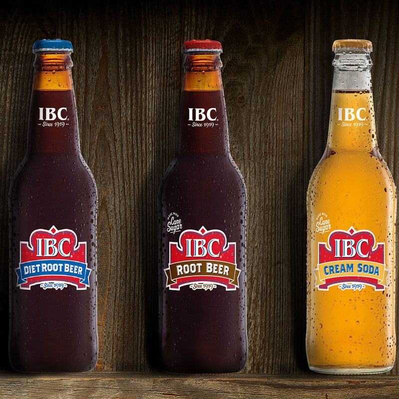 A bottle of IBC Diet Root Beer, IBC Root Beer, and IBC Cream Soda beside a red bottle cap on a wooden background.