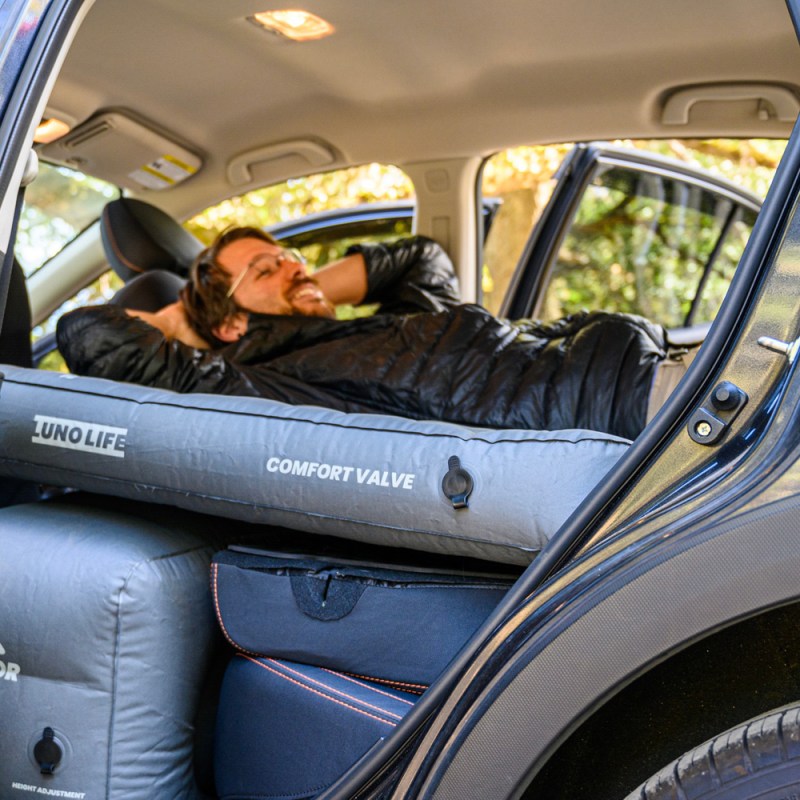A smiling bespectacled man with his hands on the back of his head is seen relaxing on a Luno air mattress in a vehicle.