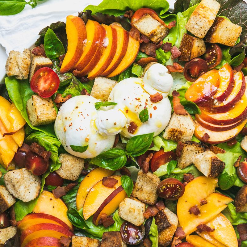 A close up of a plate of peach panzanella salad with burrata and bacon.