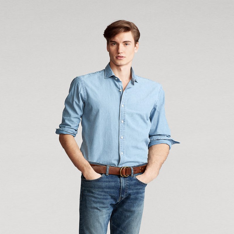 A man with his hands in his pockets is seen wearing jeans and a blue Chambray dress shirt.