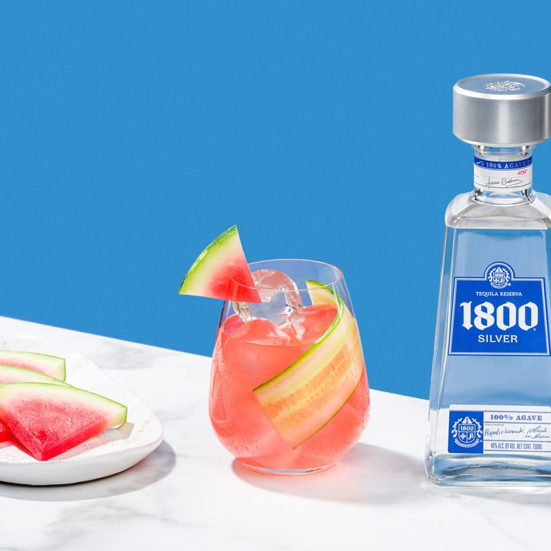 A glass of Summer Sangria in between a plate of watermelon slices and a bottle of 1800 Silver Tequila on a marble table.