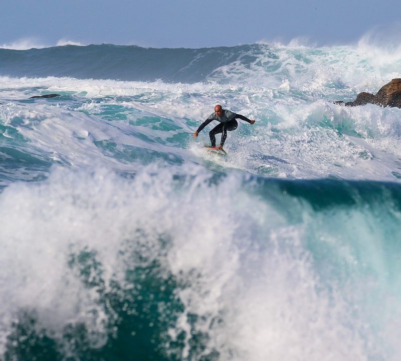 surfer in turbulent waters with jagged rocks in the background.