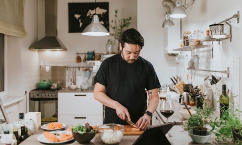 A man cuts salmon while looking at his tablet in the kitchen.