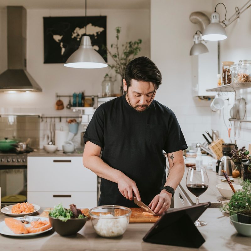 A man cuts salmon while looking at his tablet in the kitchen.