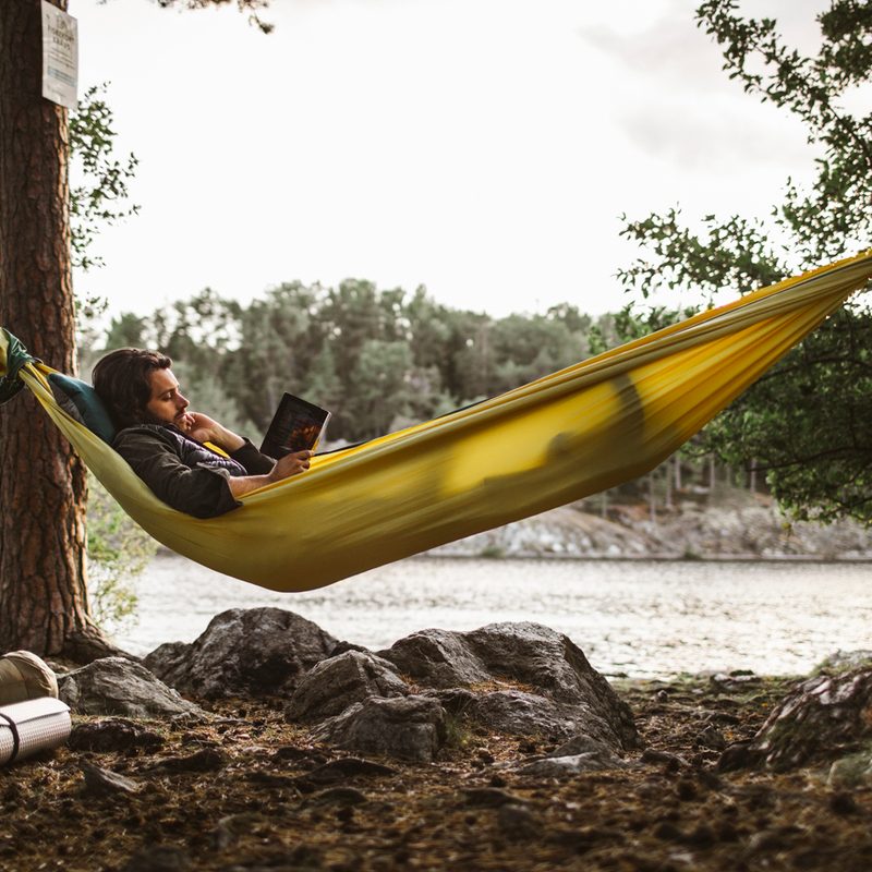 A man reading a book while lying down on a yellow hammock in the forest.