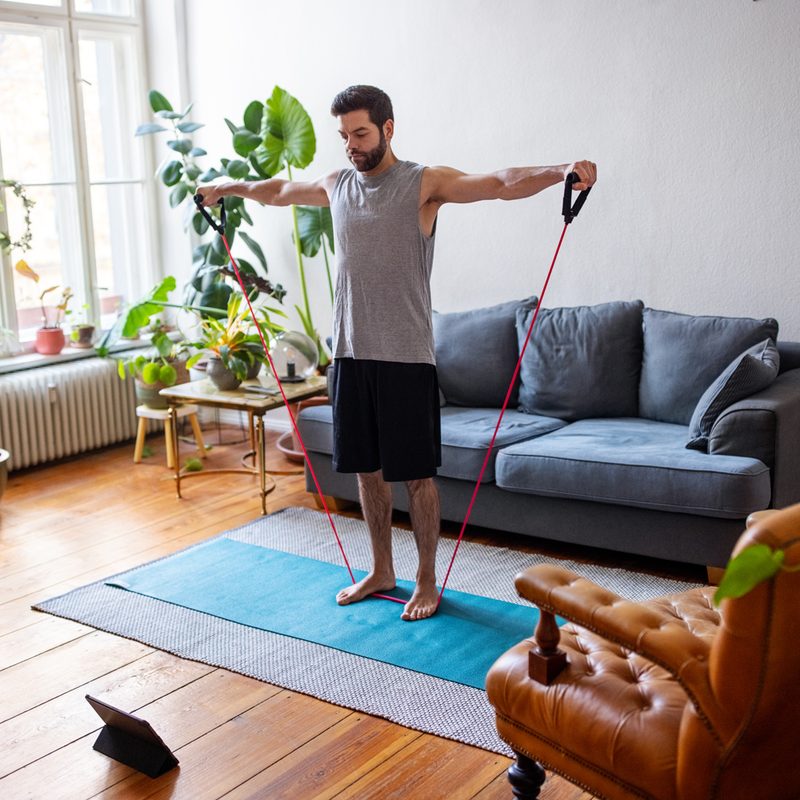 A man exercising with a stretching band while watching a video from his tablet at home.