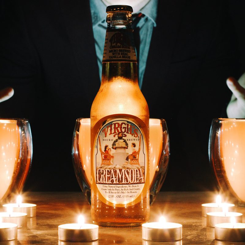 A man with his palms up, three glasses, candles, and a bottle of Virgil’s Cream Soda in the middle of a table.