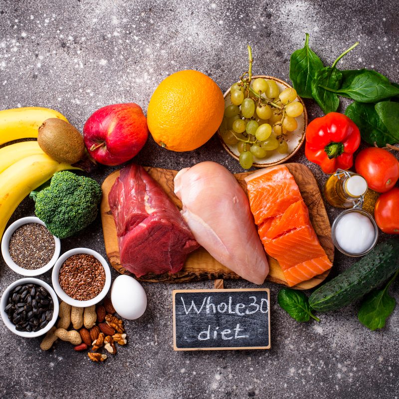An overhead shot of fruits, vegetables, unprocessed meat, nuts, and seeds with "Whole 30 diet" written on a small blackboard.