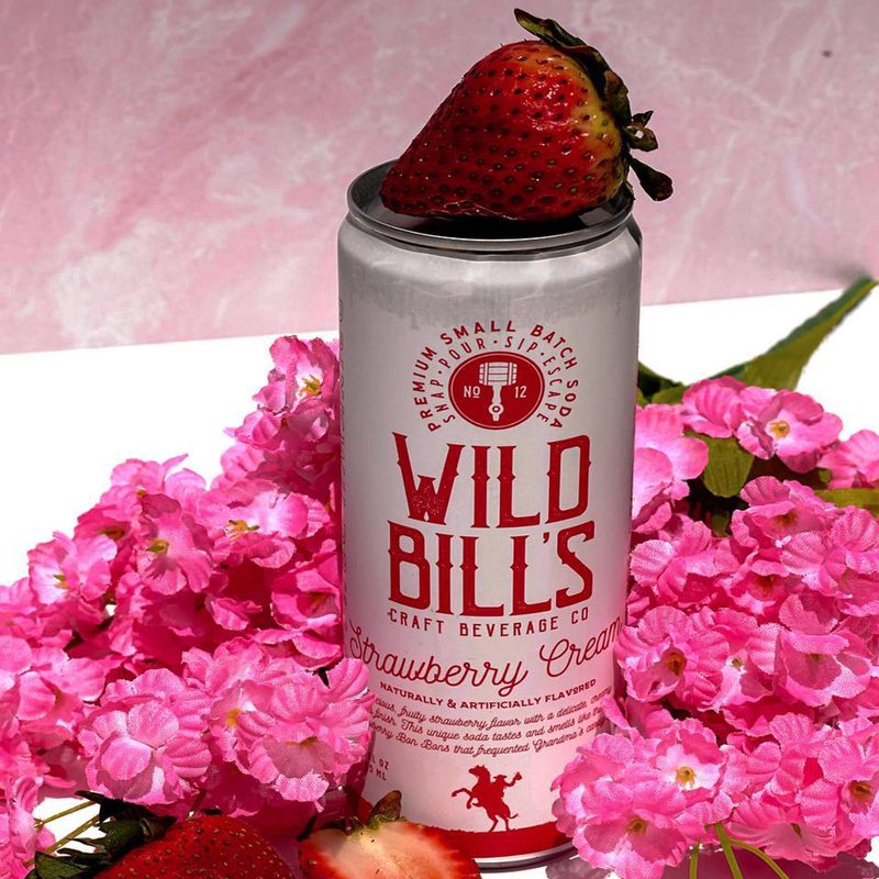 A strawberry on top of a can of Wild Bill's Strawberry Cream Soda surrounded by strawberry slices and pink flowers on a white table.