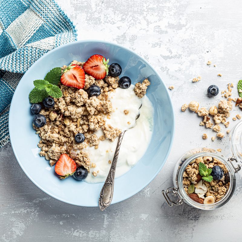 A blue towel, a bowl of yogurt with granola, blueberries, strawberries, and a mint leaf, and a jar of granola on a table.
