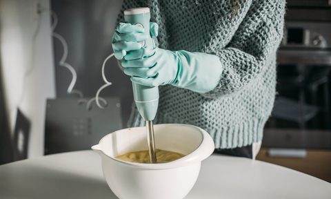 hands with protective gloves using a hand blender on a white bowl.