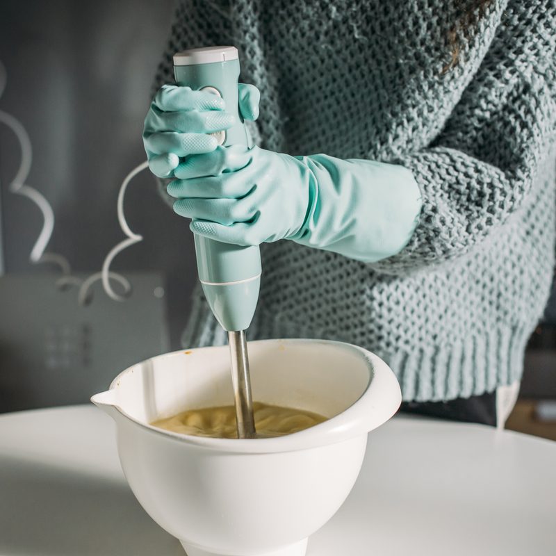 hands with protective gloves using a hand blender on a white bowl.