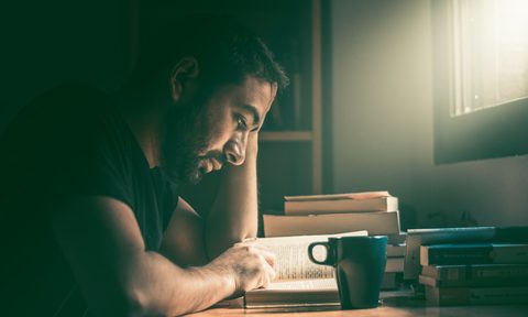 Young man reading a novel with a cup of coffee next to stack of books.