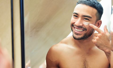 Shot of a young man looking in the mirror while applying moisturizer to his face.