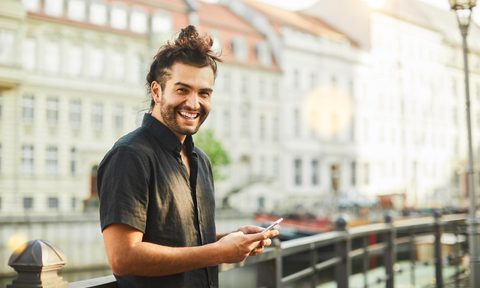 A smiling man in front of a landmark is seen wearing a black short-sleeve shirt and holding a phone.