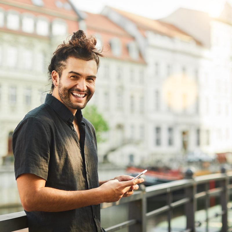 A smiling man in front of a landmark is seen holding a phone and wearing a black short-sleeve shirt.
