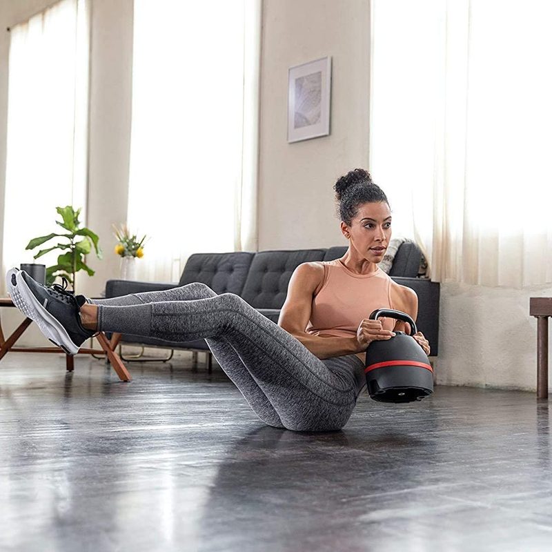 Woman using the Bowflex SelectTech Kettlebell in a twist weight pilates exercise while seated on the floor of her living room.