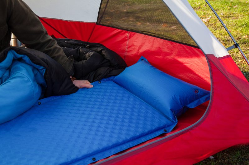 A man gets his tent and sleeping bag ready at a campground by inflating and setting up his blue blow-up mattress pad to put for under his sleeping bag.