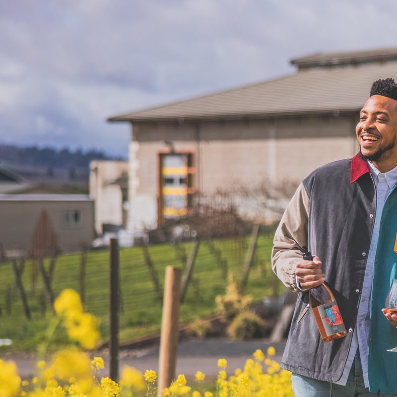 CJ McCollum smiling while holding a wine bottle and glass at the vineyard.
