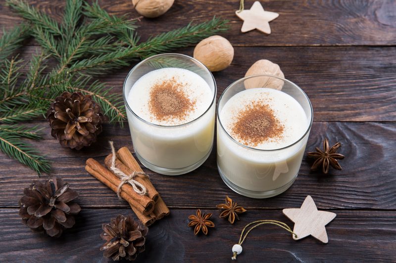 Eggnog with cinnamon in glasses on wooden background with Christmas decor, homemade traditional festive drink for Christmas time.