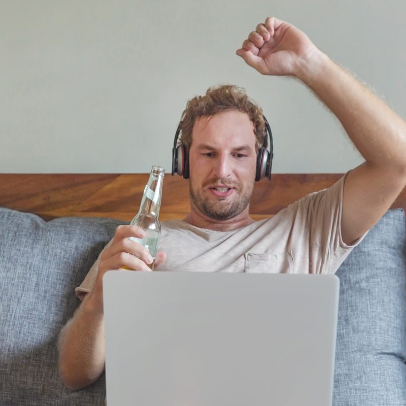 A man with beer bottle raising his arm while watching an online performance from his laptop.