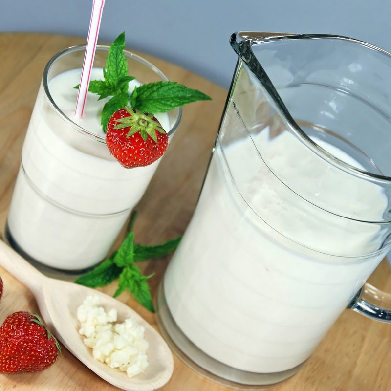 A jug and glass of kefir with strawberry, a spoon filled with grains of kefir, two strawberries, and mint leaf on a round wooden table.
