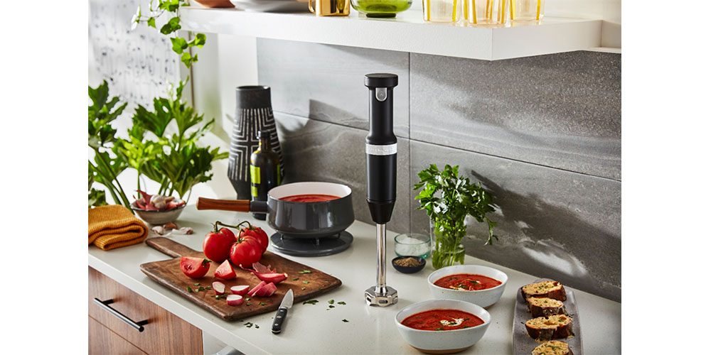KitchenAid Cordless Variable Speed Hand Blender placed on a kitchen countertop surrounded by food.