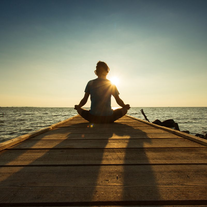 Young man sitting and meditating on a pontoon by a lake at the sunrise.