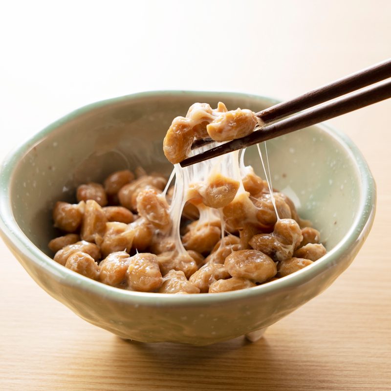 A serving of Natto is lifted with chopsticks from a bowl on a wooden table.