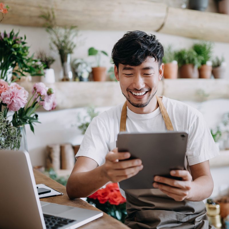 A male florist happily using his iPad in his flower shop.