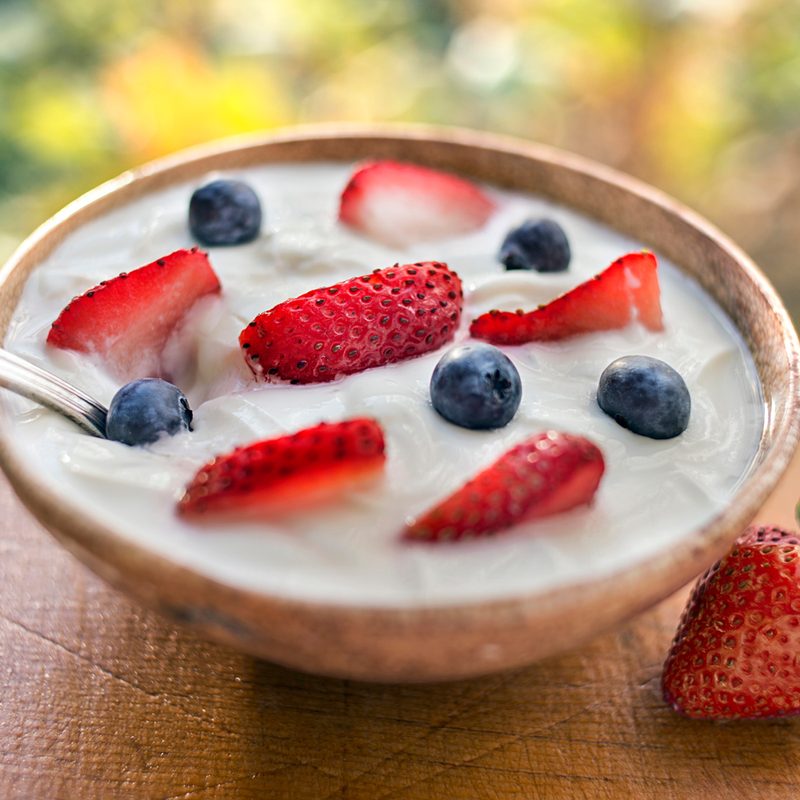 A spoon dipped into a bowl of yogurt with strawberry slices and blueberries beside a strawberry.