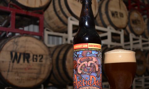 A bottle and glass of Midnight Sun Arctic Devil with barrels as the backdrop.
