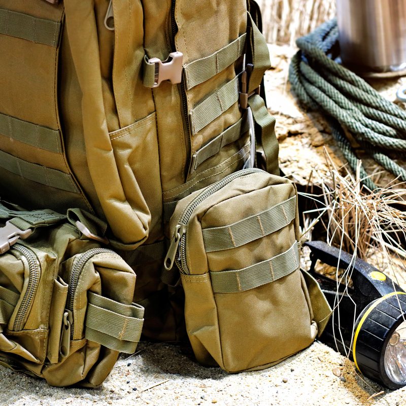 A tactical backpack beside a flashlight and rope on sand.